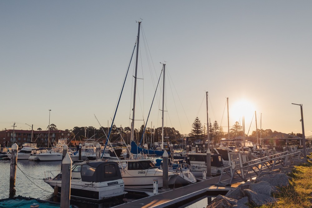 [Image Description: white boats in a marina, rocks and ramps surrounding the area, blue sky above and sun setting in the middle right side of image.]