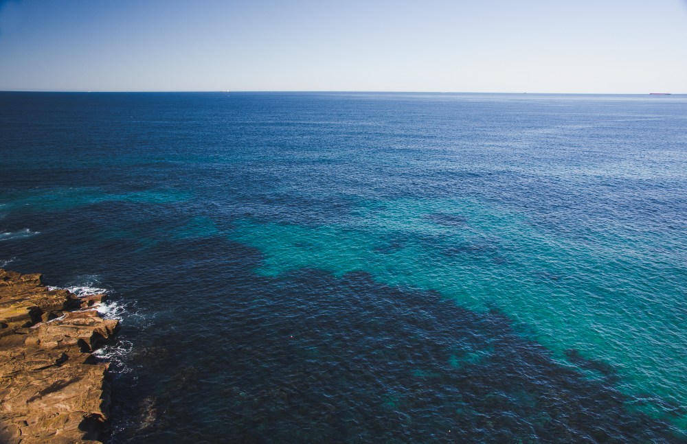 [Image Description: Blue and teal deep sea bed, brown rock cliff to the left of the image. Looking out to the ocean on the horizon.]