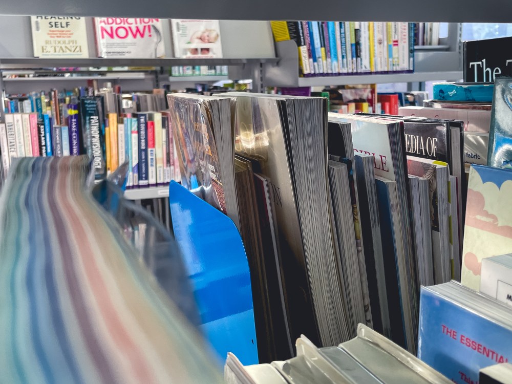 [Image Description: Colourful library books arranged on shelves, image is looking through the shelves to the next aisles.]