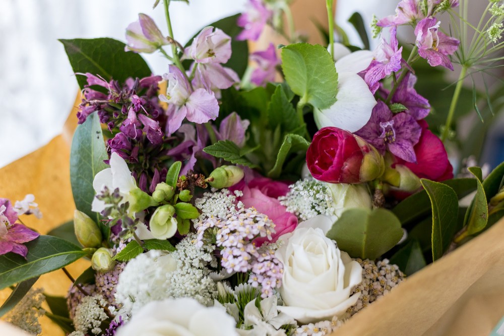 [Image Description: A bunch of mixed flowers, wrapped in brown paper with a white lace curtain in the background.]