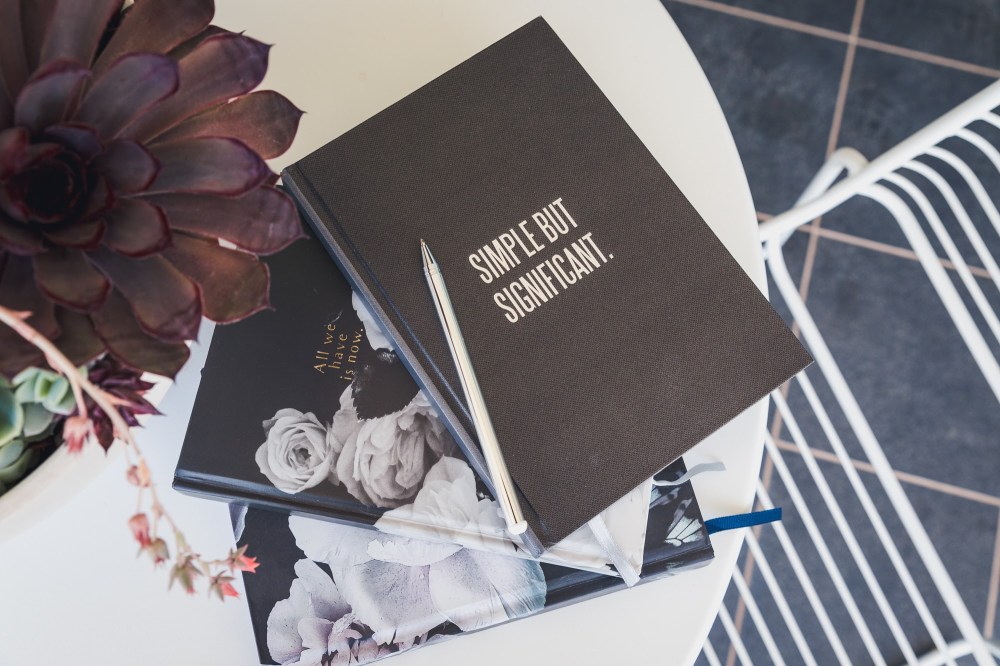 [Image Description: Black and dark floral journal books stacked with silver pen on top, on white round table and a white pot full of succulent plants, with a white wire chair on dark grey tiles.]