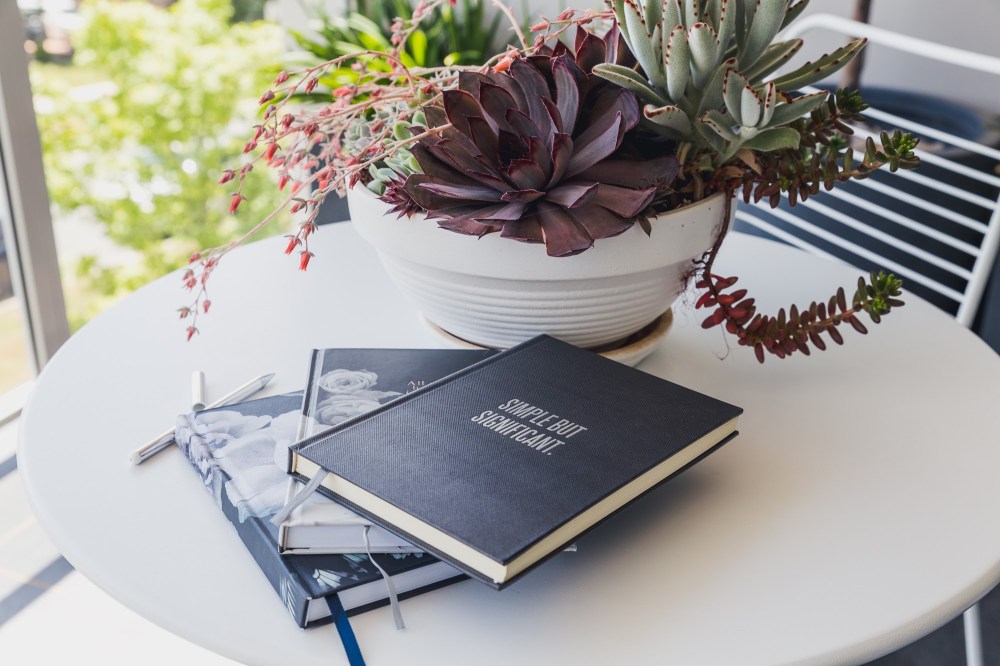 [Image Description: Black and dark floral journal books stacked on a white round table and a white pot full of succulent plants, with a white wire chair and a green tree in the background.]