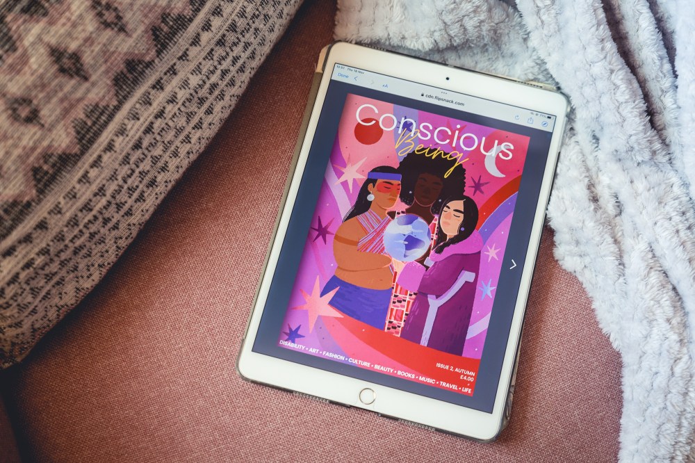 [Image Description: A white iPad with Conscious Being Issue 2 magazine open on the screen. Pink and purple cover magazine with three Indigenous women holding a sphere with stars around them. The iPad is on a pink armchair with a grey fleece blanket and a pink pattern cushion next to it.]