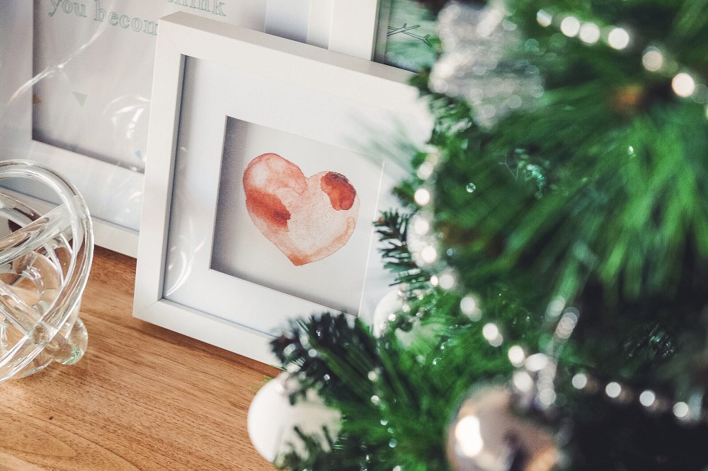 [Image Description: Blurry green Christmas tree with silver decorations, and focus on white photo frames with a red watercolour heart on a timber table.]