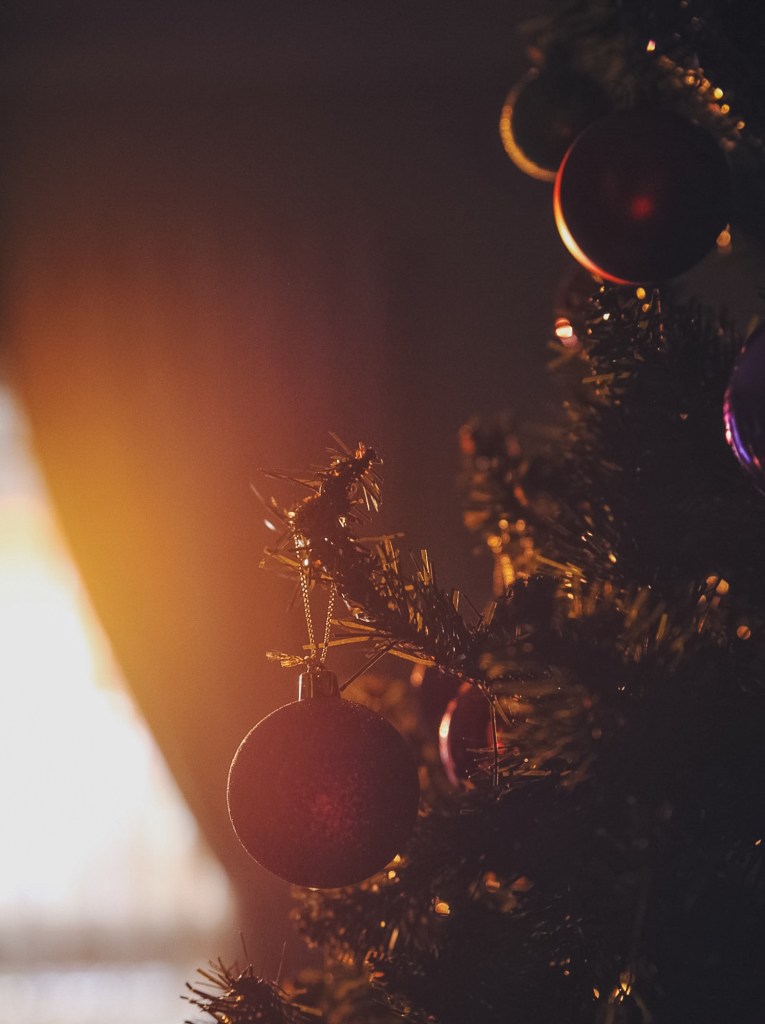 [Image Description: Golden light shining on a dark baubles and a green pine Christmas tree, with a curtain in the background.]
