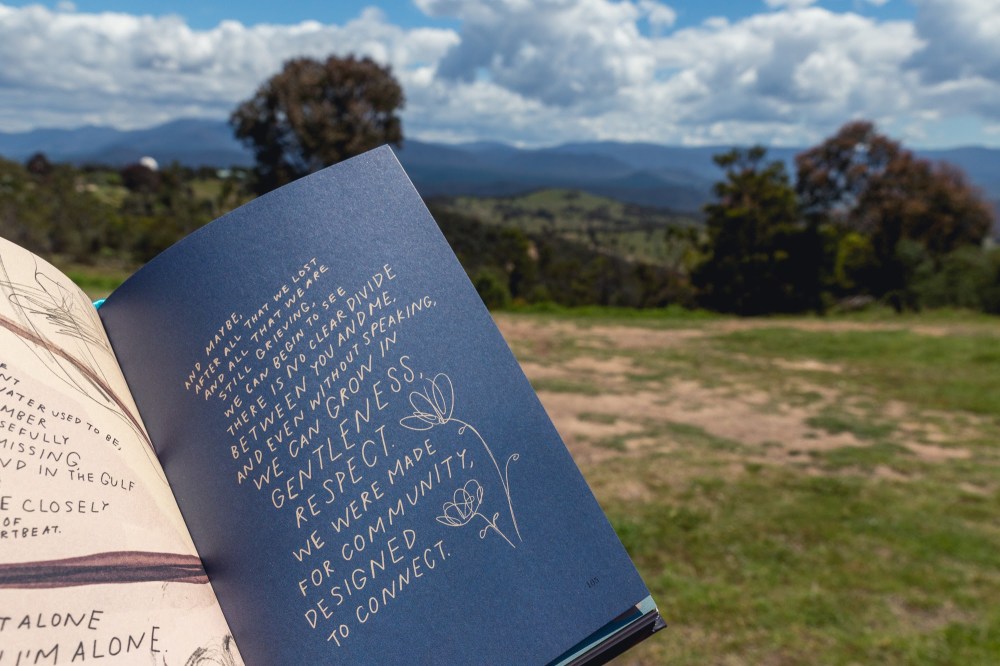 [Image Description: Book page held up open with grass, bush, clouds and mountains in the background. On the navy page is cream illustration of a flower and poetry by Morgan Harper Nichols.]