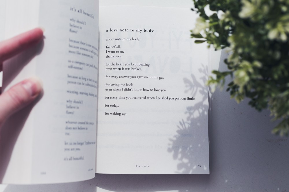 [Image description: book opened to a page of poetry, with two pink fingers holding open the page, a green plant to the right and a white table underneath. The poetry book is Heart Talk by Cleo Wade, and is open to a poem called ‘a love note to my body’.]