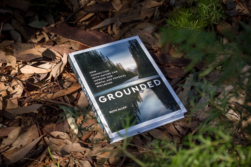 [Image Description: The book is Grounded by Ruth Allen, and has a nature scene of a lake, pine trees and mountains on the cover with large white font saying 'Grounded: How connection with nature can improve out mental and physical wellbeing'. It is placed bush litter, and is in part light and shade, with greenery around it out of focus. ]
