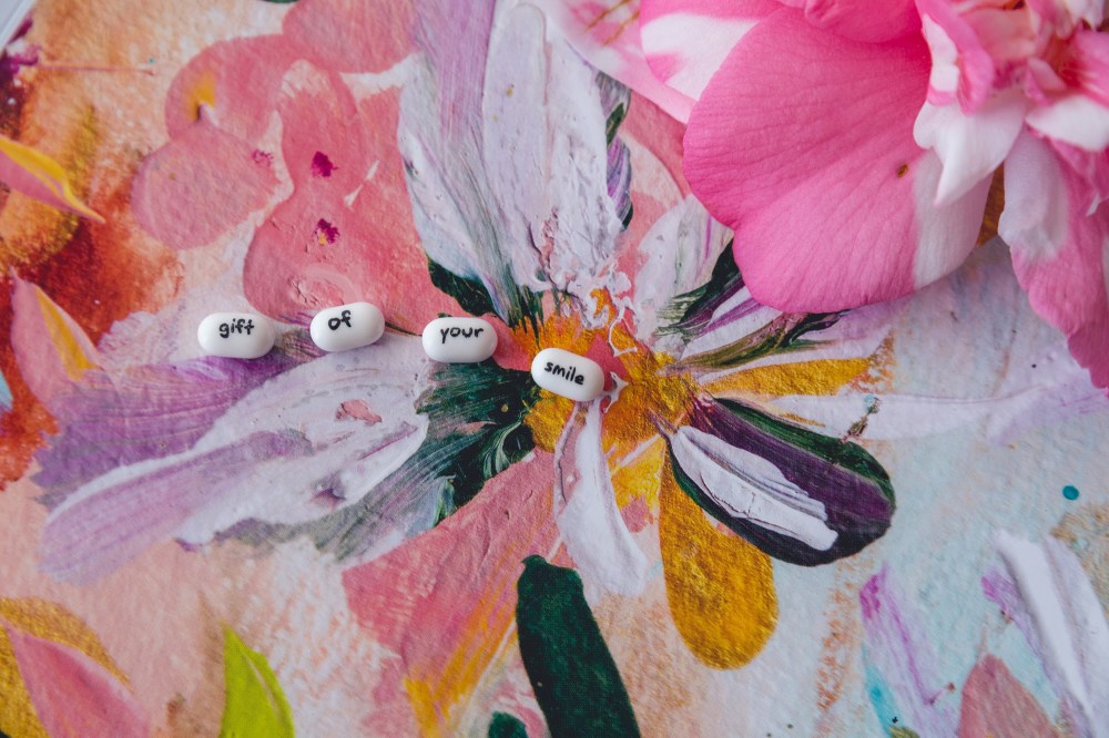 [Image Description: Four white TicTacs on a colourful pink artwork with a pink camellia flower in the top right corner. There is one word on each TicTac that reads "gift of your smile".]