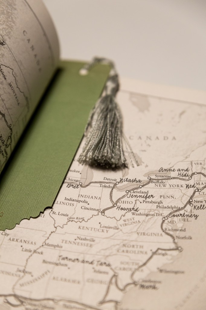 [Image Description: Book open to a map of Northern America with a journey line drawn with people's names at various locations. A green bookmark with a sage green tassel is in the spine of the book page. The book is Between Two Kingdoms by Suleika Jaouad.]