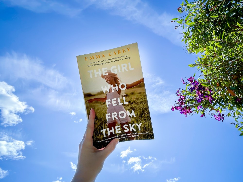 [Image description: Book held up above with the sky with wispy clouds and a green bush with purple flowers. The book is The Girl Who Fell From The Sky by Emma Carey. It has a girl in a white mini dress in a field of grass, with large white text.]