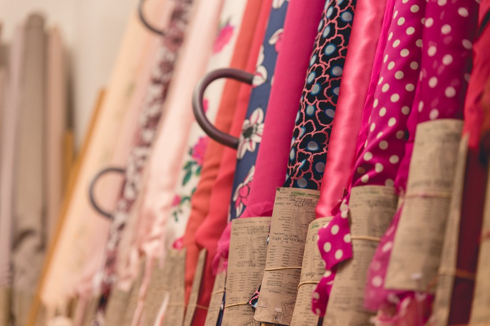 [Image description: cream and pink patterned rolls of fabric lined up on a stand, with brown paper description cards secured on each roll with rubber bands.]