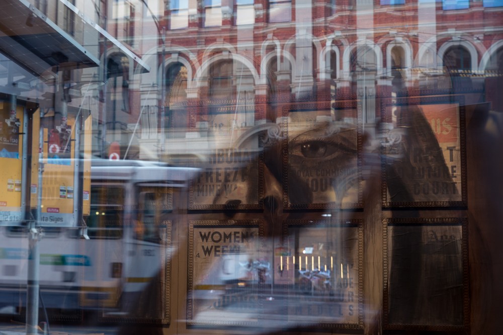 [Image description: a multi layered image of a mural of a woman's face on a wall of vintage posters, with glass reflections of the street buildings and traffic. The mural and art installation is by the artist RONE.]