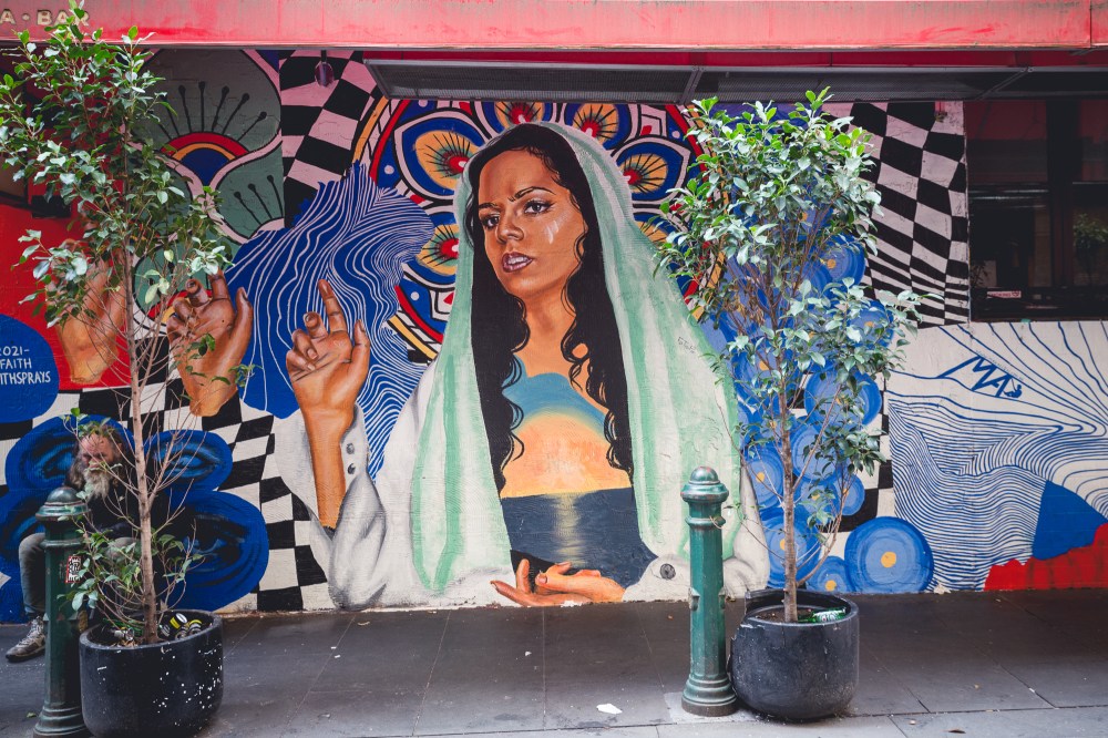 [Image description: painted mural of a woman with a scarf over her head, with one hand raised to ask a question. The background of the mural is colour patterns and there are two potted trees on either side on the street in front of the mural.]