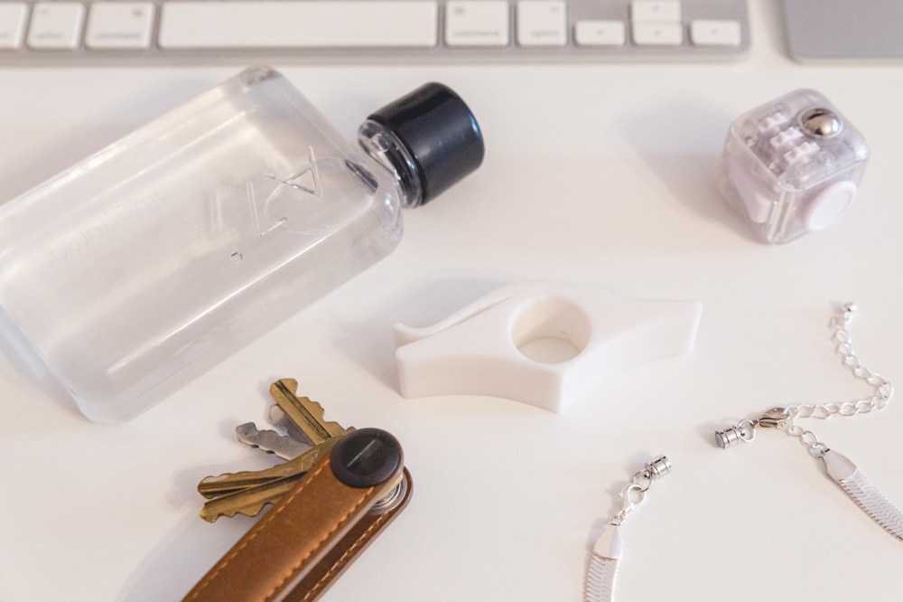 [Image Description: Flat lay image on a white surface with a small rectangular water bottle with a black lid, a silver and white computer keyboard, a fidget toy, a book page opener, silver necklace clasps and a tan leather Orbitkey organiser with gold and silver keys splayed out.]